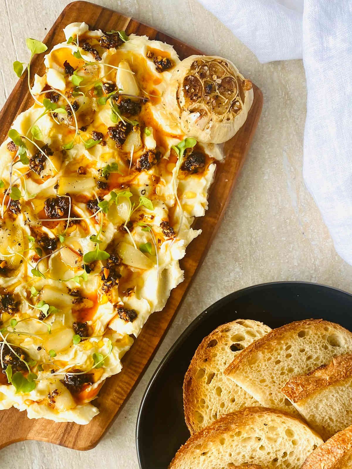 butter board with butter, garlic, salt, chili crisp oil, honey and microgreens with a whole roasted garlic in the corner and a plate of crusty toasted french bread.