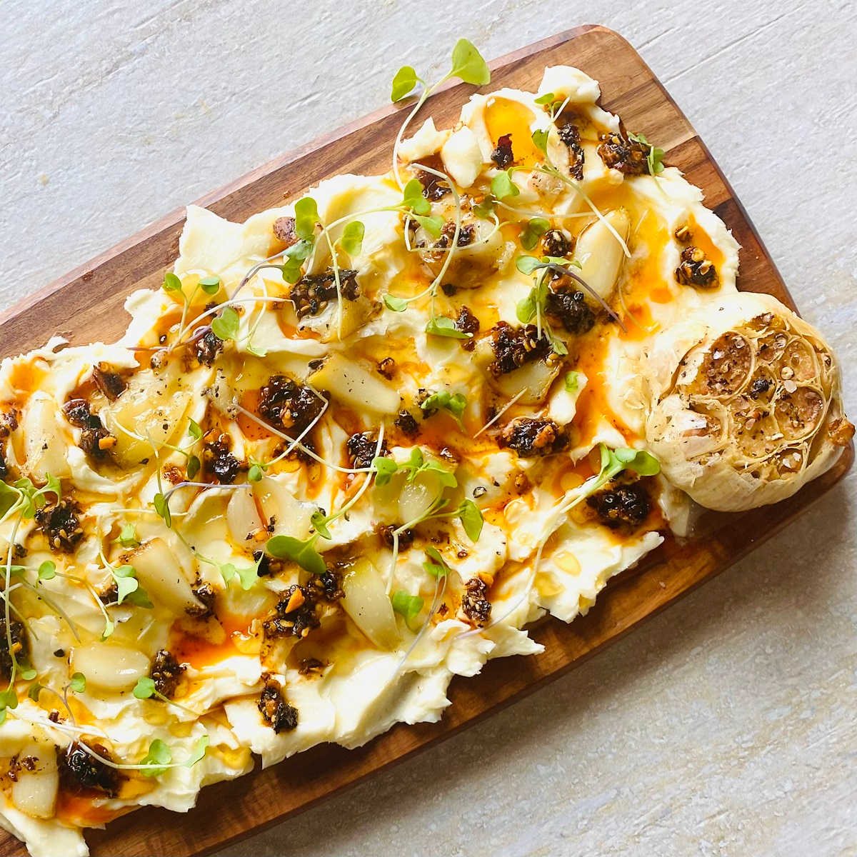 butter board recipe with butter, garlic, salt, chili crisp oil, honey and microgreens with a whole roasted garlic in the corner.