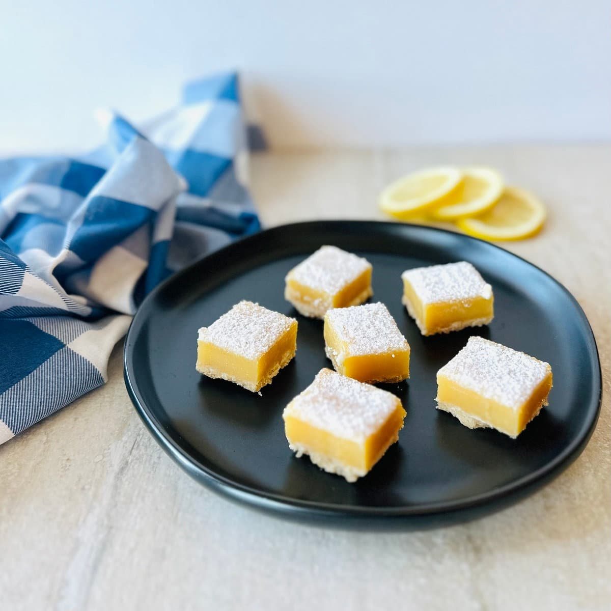 lemon squares with graham cracker crust on black plate with sliced lemons and a blue napkin.
