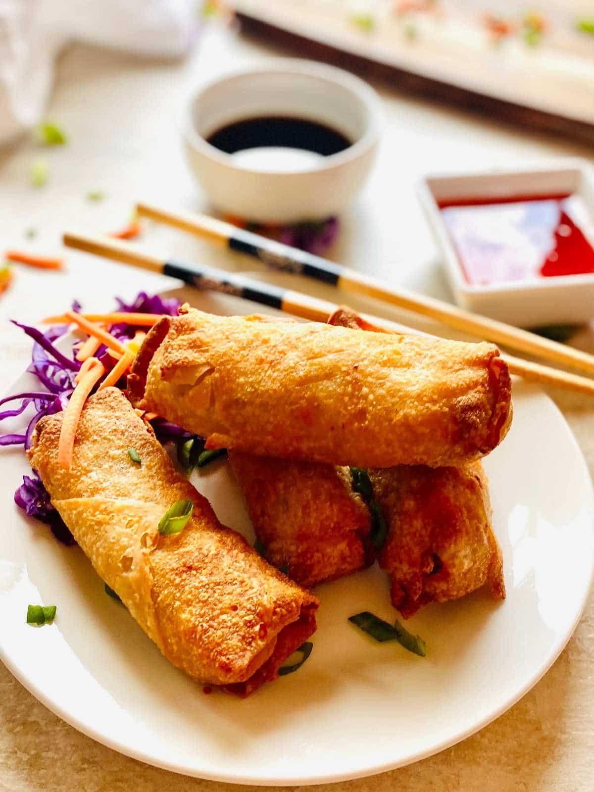 air fryer frozen egg rolls on a plate next to chopstick, soy sauce and thai chili dipping sauce