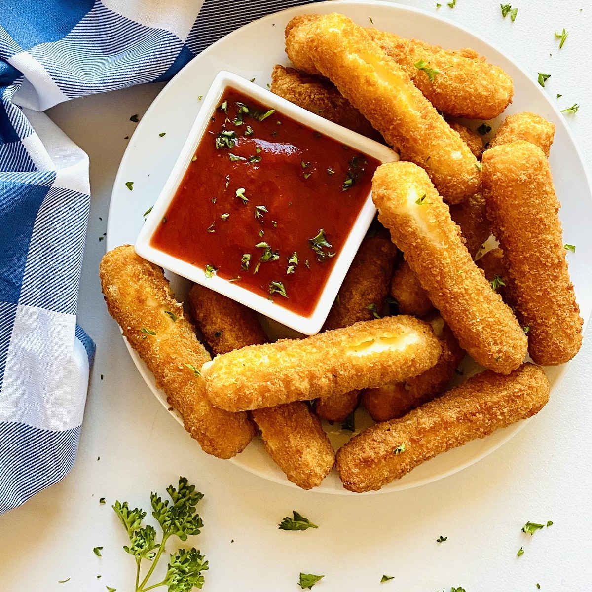 mozzarella sticks on a white pate next to a side dish or marinara sauce and a blue napkin