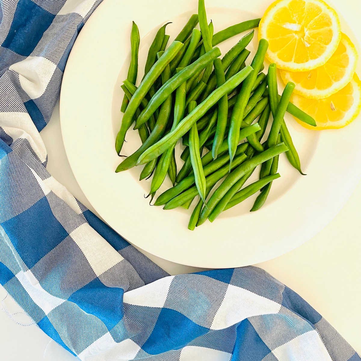 green beans on a plate next to lemon slices and a blue checkered cloth napkin