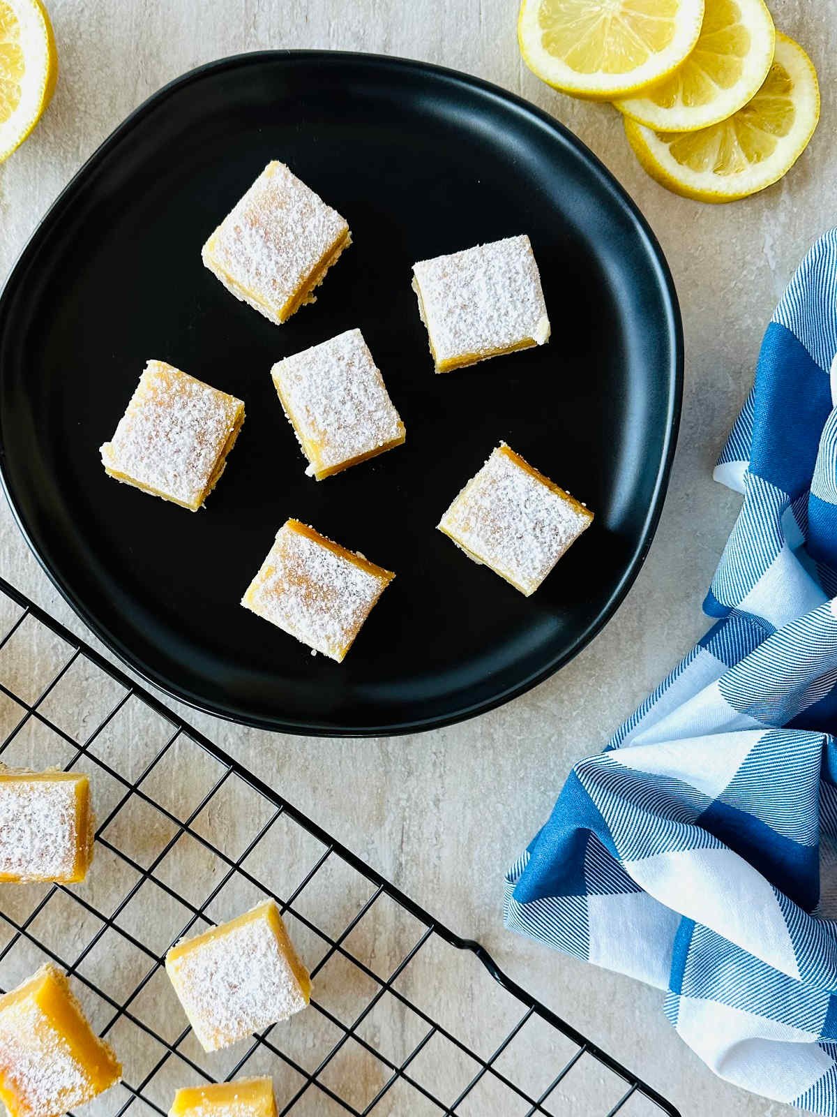 lemon bars with graham cracker crust on black plate with sliced lemons and a blue napkin.