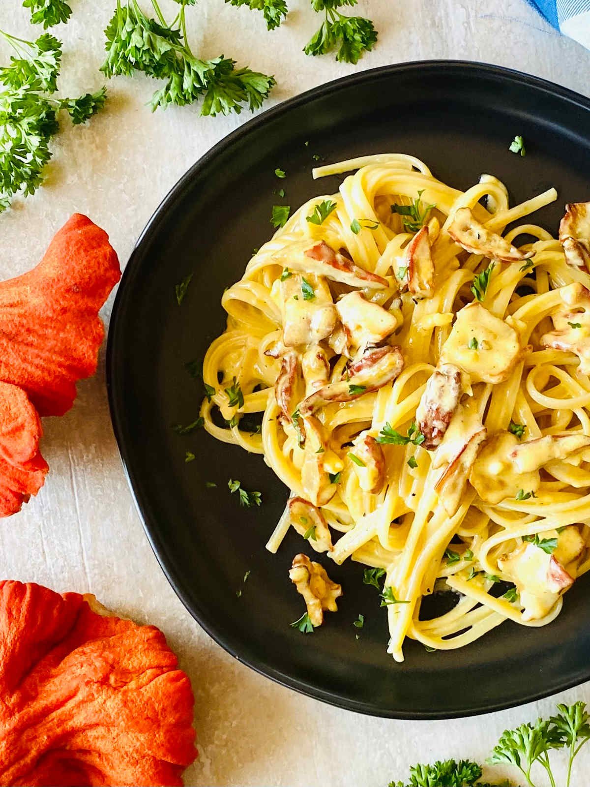 lobster mushroom pasta with cream sauce on a black plate next to fresh lobster mushrooms, parsley and a blue napkin