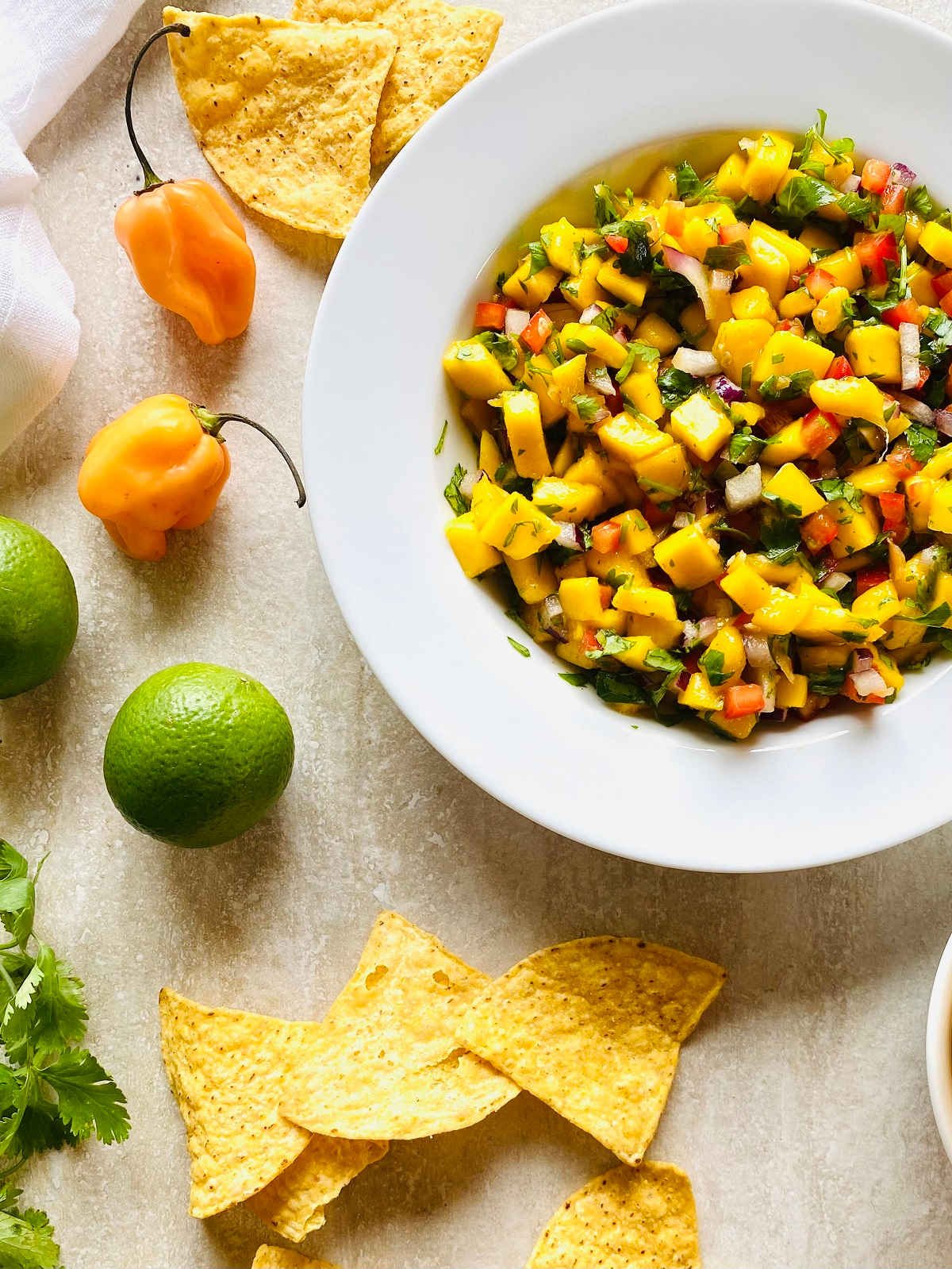 mango habanero salsa in a bowl surrounded by chips, habanero peppers, limes and cilantro