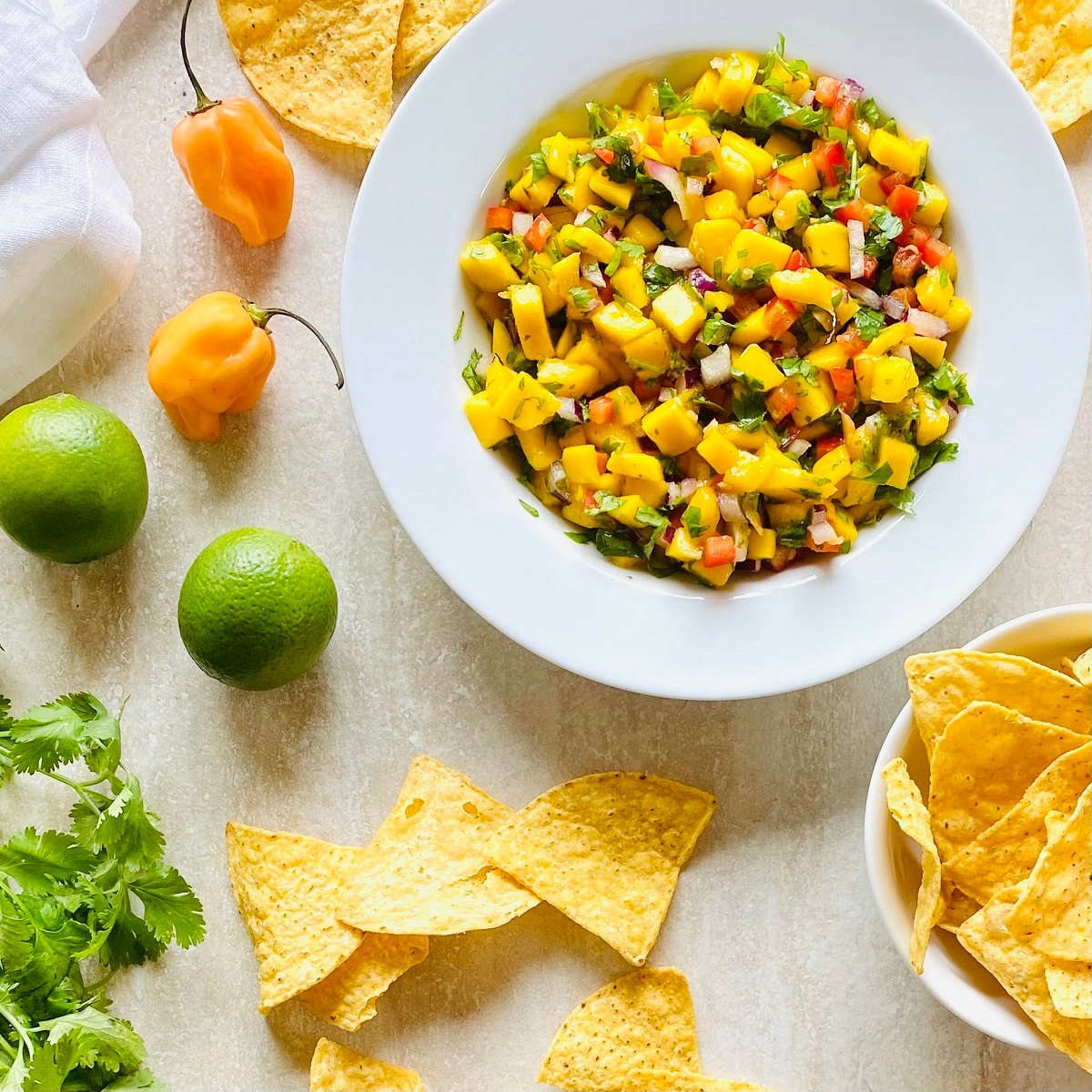mango habanero salsa in a bowl surrounded by chips, habanero peppers, limes and cilantro