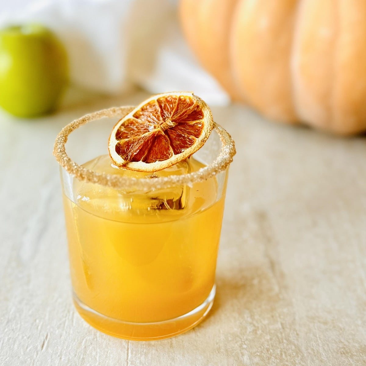 Maple Apple Cider Old Fashioned Recipe in glass with cinnamon sugar rim and orange slice garnish, with fairytale pumpkin in background.