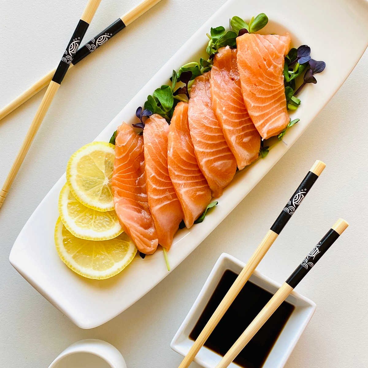 salmon sashimi on a bed of microgreens garnished with lemon next to soy sauce and chopsticks