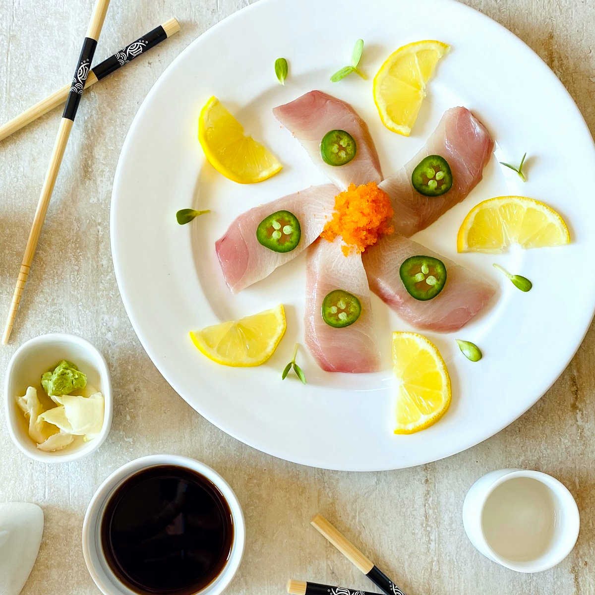 yellowtail sashimi slices on white plate topped with jalapeno slices and surrounded by lemon slices, soy sauce and chopsticks