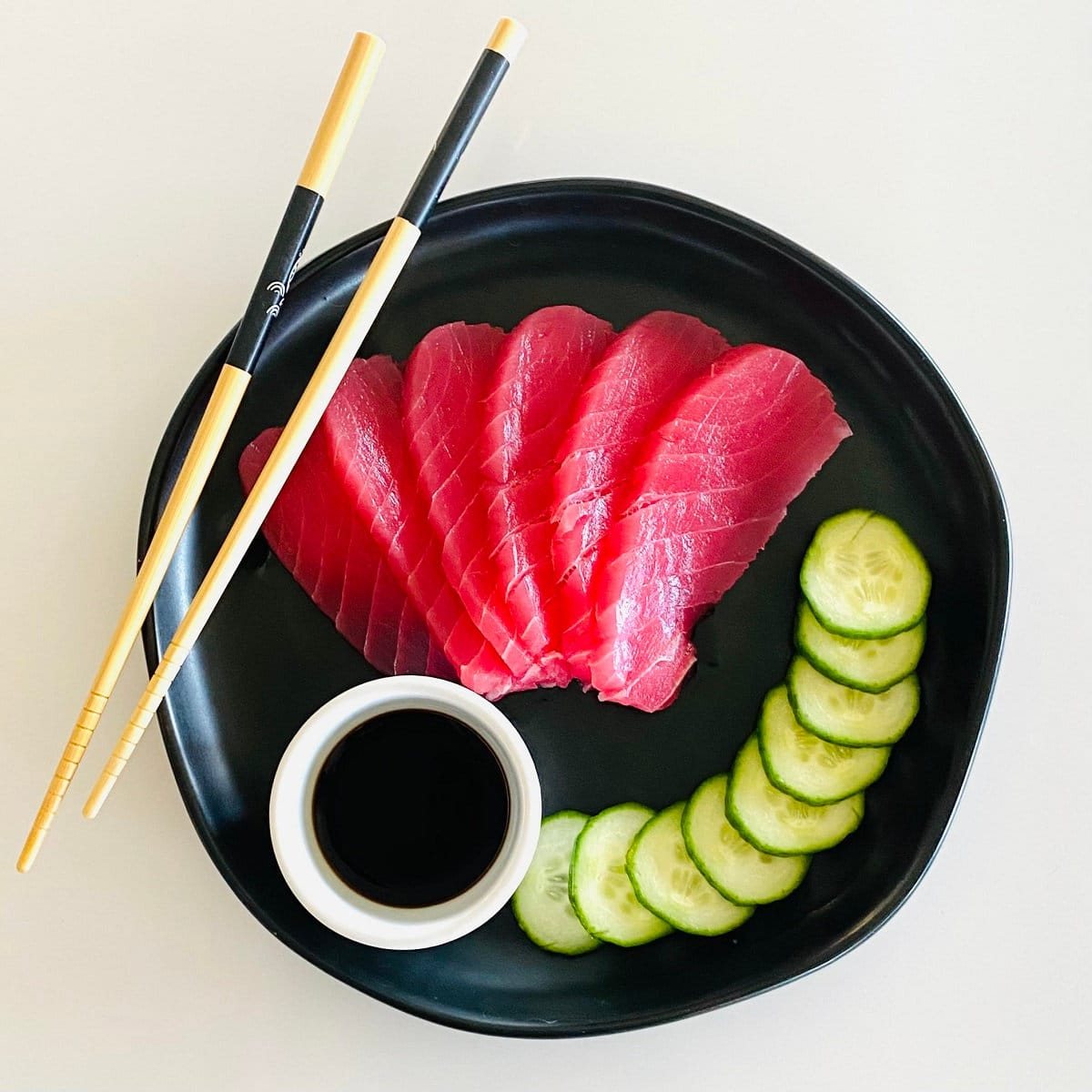 tuna sashimi recipe fully prepared on plate with soy sauce, sliced cucumbers and chopsticks.