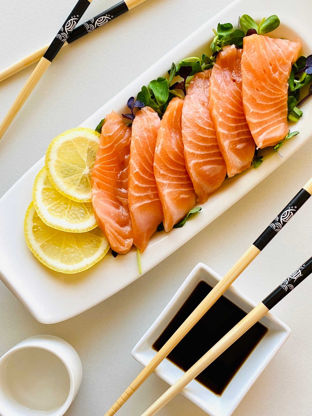 salmon sashimi on a bed of microgreens garnished with lemon next to soy sauce and chopsticks