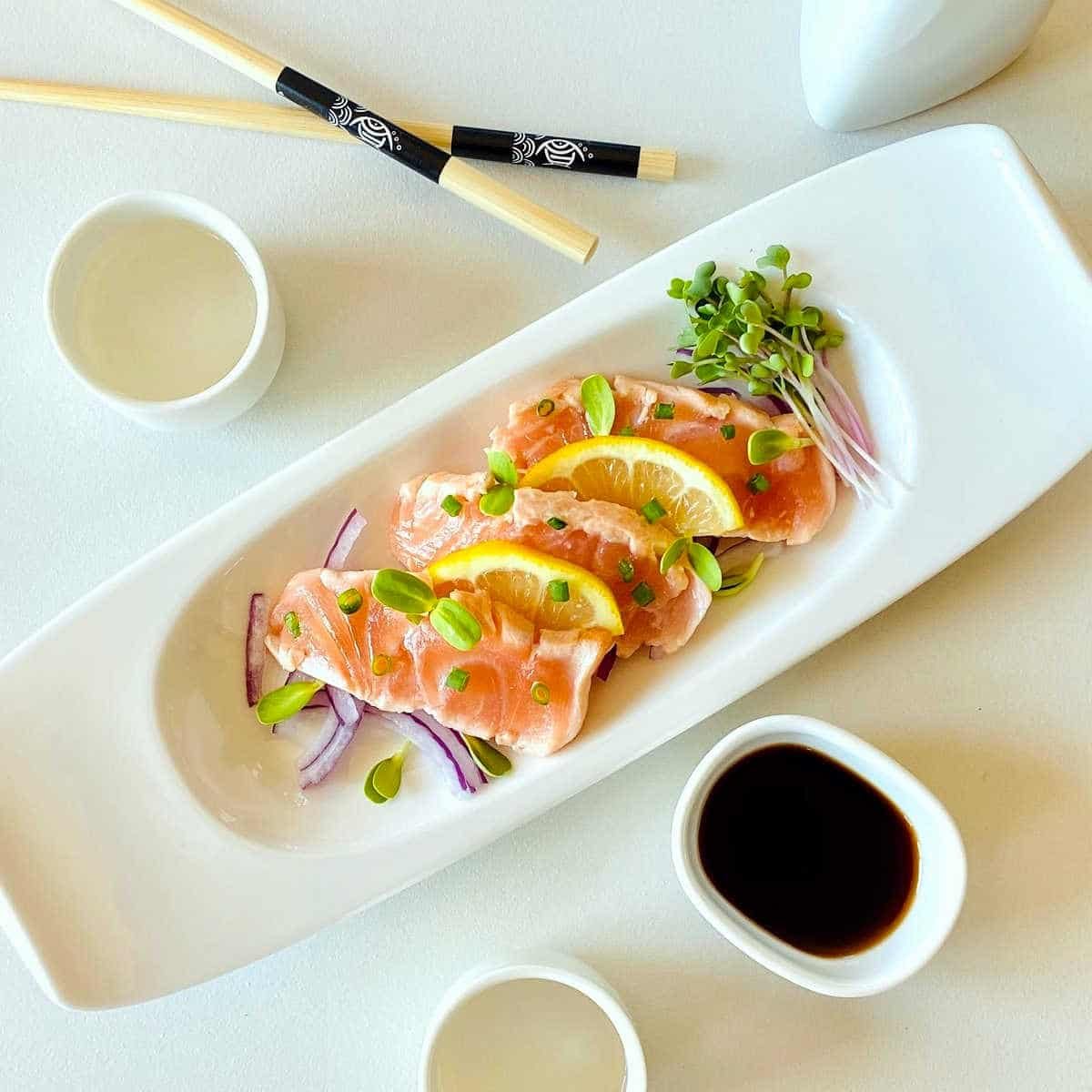 salmon tataki on plate garnished with lemon onion and sprouts next to chopsticks and ponzu