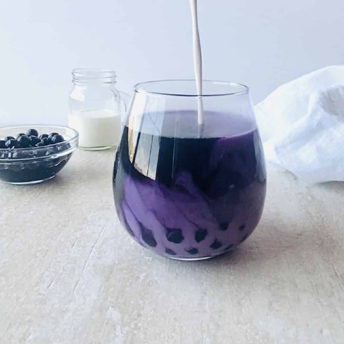 milk being poured into taro milk tea with boba in the background