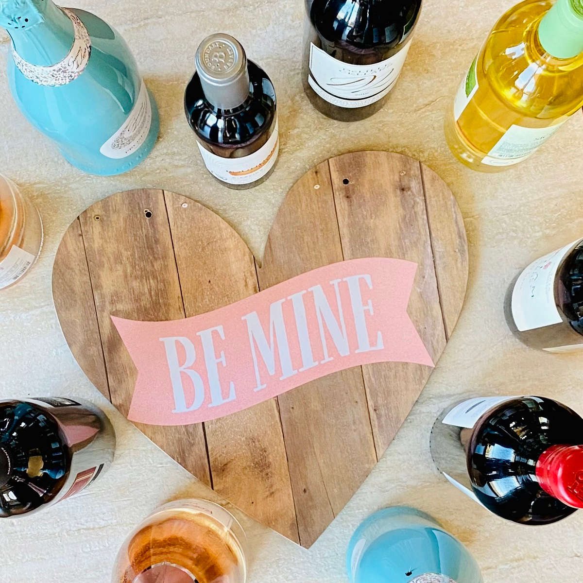 a heart board that says "be mine" surrounded by different bottles of wine