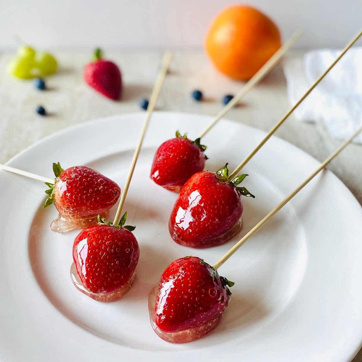 tanghulu strawberries on white plate with orange strawberry and grapes in background