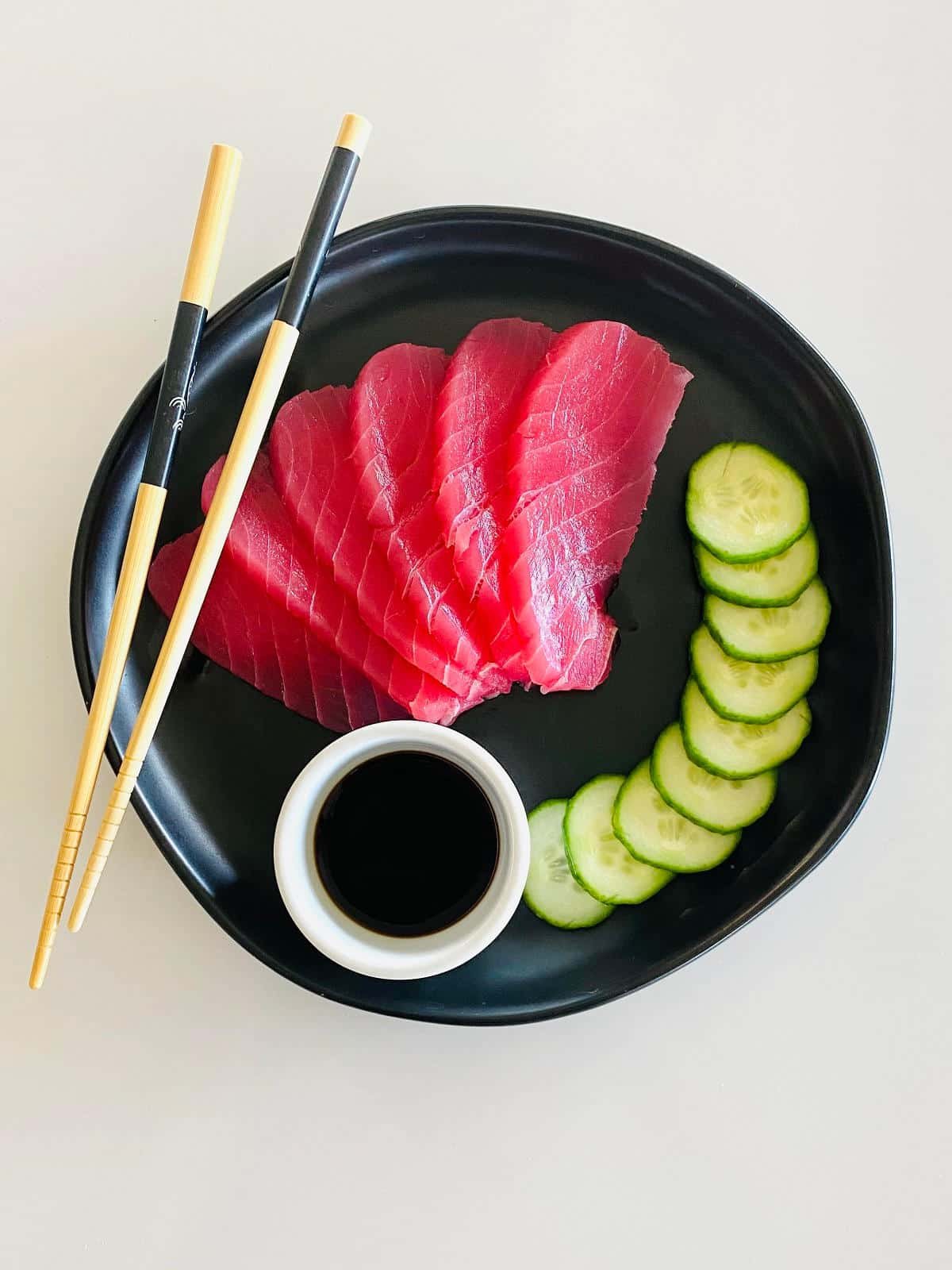 ahi tuna sashimi on plate with soy sauce, sliced cucumbers and chopsticks.