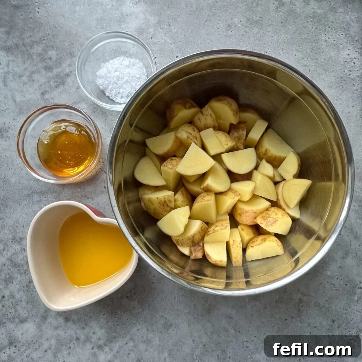 Golden Honey Glazed Potatoes 4 A metal bowl of chopped potatoes, a small bowl of melted butter, a small bowl of honey, and a small bowl of salt on a gray surface.