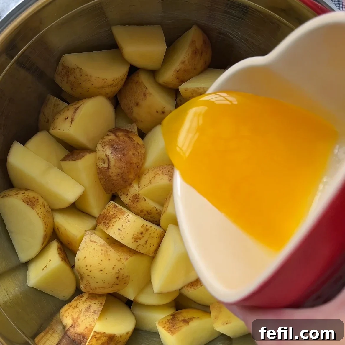 Golden Honey Glazed Potatoes 6 A hand pours melted butter from a red cup onto chopped, unpeeled potatoes in a metal pot, preparing them for delicious Honey Roasted Potatoes.