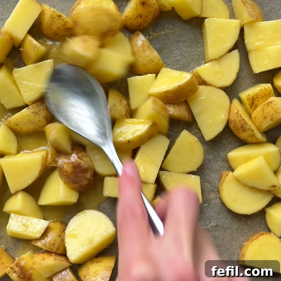 Golden Honey Glazed Potatoes 7 A hand uses a spoon to spread chopped, unpeeled potatoes coated in butter and honey onto a baking sheet, preparing them for honey roasted potatoes.