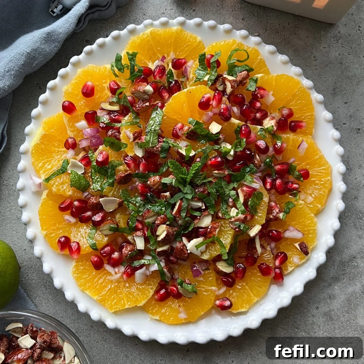 A plate of sliced oranges topped with glistening pomegranate seeds, crunchy chopped nuts, fresh mint leaves, and delicate red onion slices, all arranged artfully on a sleek gray surface.