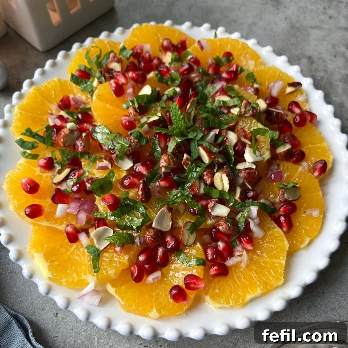 A close-up side view of a beautifully composed plate of sliced oranges, garnished with bright red pomegranate seeds, chopped almonds, finely diced red onions, and fresh mint sprigs, presented on an elegant white dish.