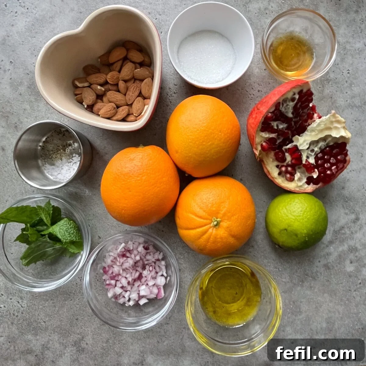 A flat lay arrangement showcasing the fresh ingredients for the orange salad: whole almonds, granulated sugar, whole oranges, a cut pomegranate, a fresh lime, sprigs of fresh mint, finely chopped red onion, a bottle of olive oil, a pinch of sea salt, and a drizzle of honey, all neatly presented in small bowls on a gray surface.