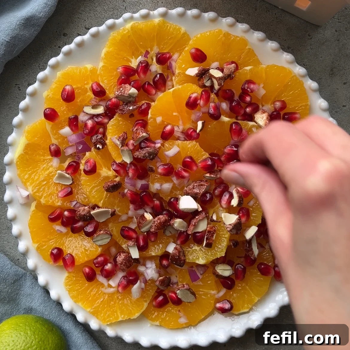 A hand gracefully sprinkles an assortment of colorful toppings, including glistening pomegranate seeds, finely chopped nuts, and delicate diced red onions, over a vibrant plate of sliced oranges.