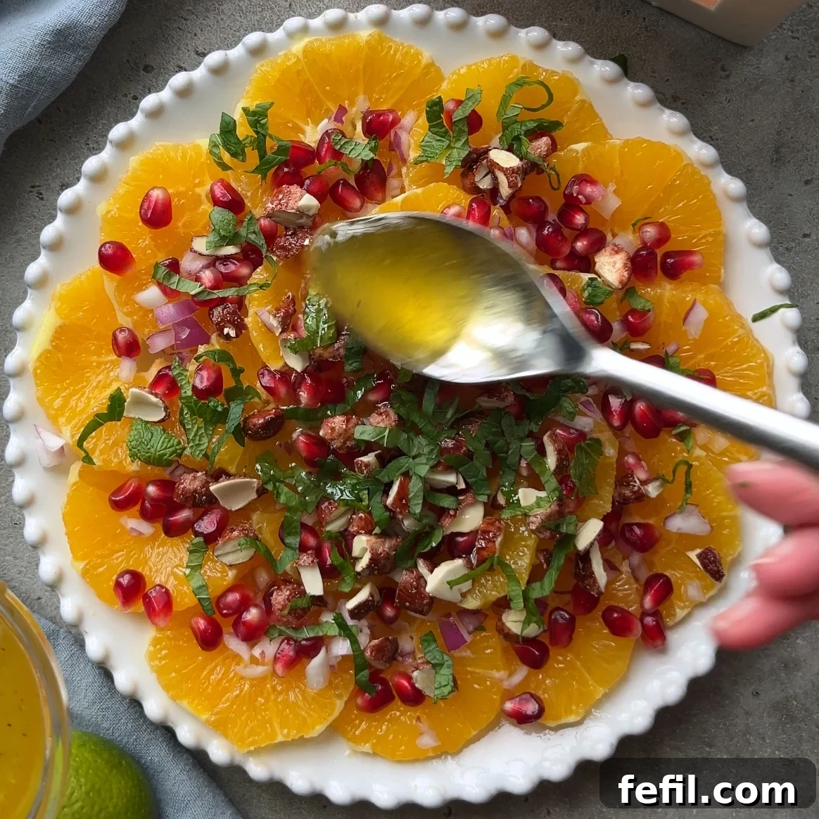 A hand skillfully drizzles a light, glossy dressing over a captivating plate of sliced oranges, which are beautifully topped with vivid pomegranate seeds, crunchy chopped nuts, fresh mint leaves, and finely diced red onion.