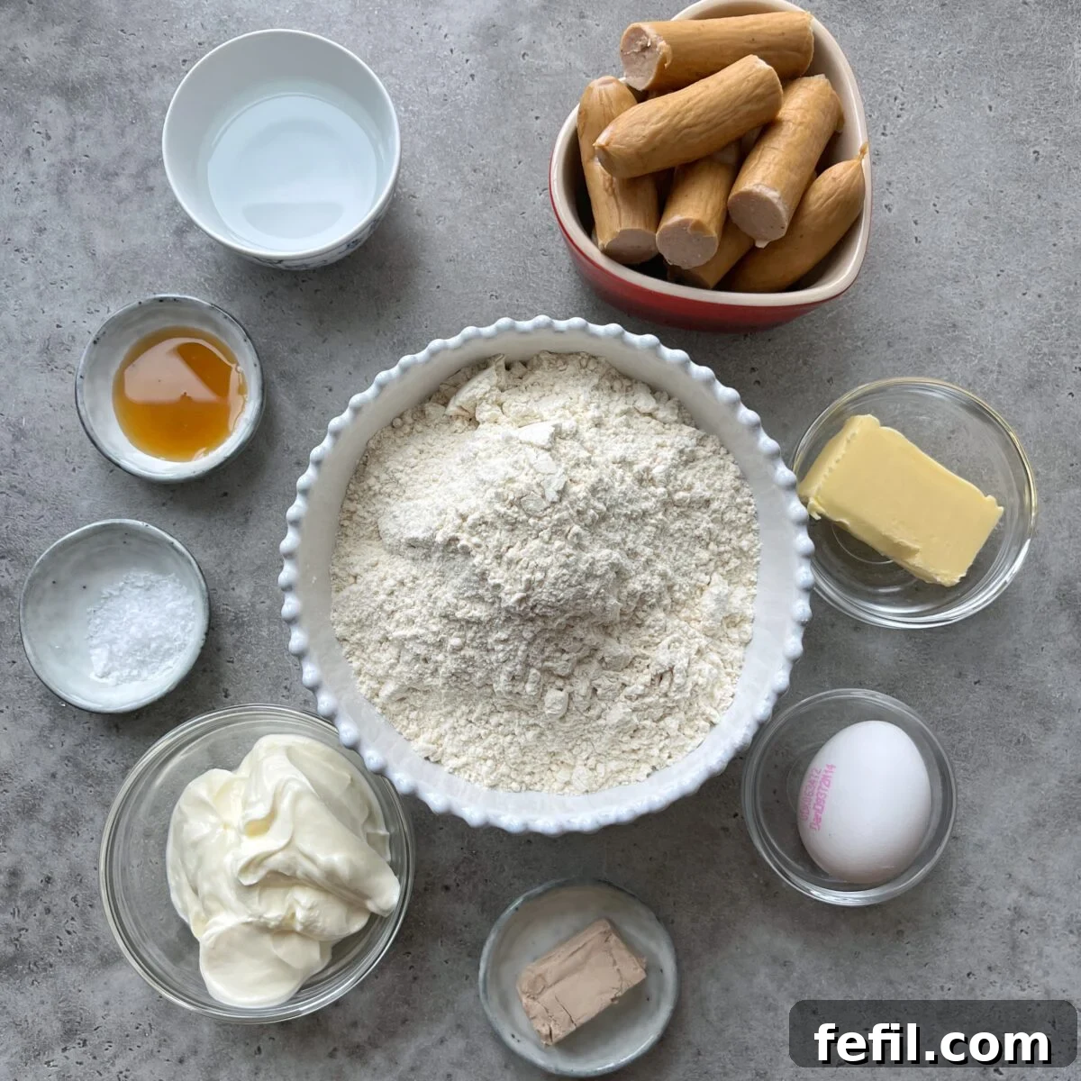 Assorted baking ingredients in bowls on a grey surface, including flour, butter, maple syrup, sour cream, baking powder, yeast, water, egg, and several uncooked sausages.