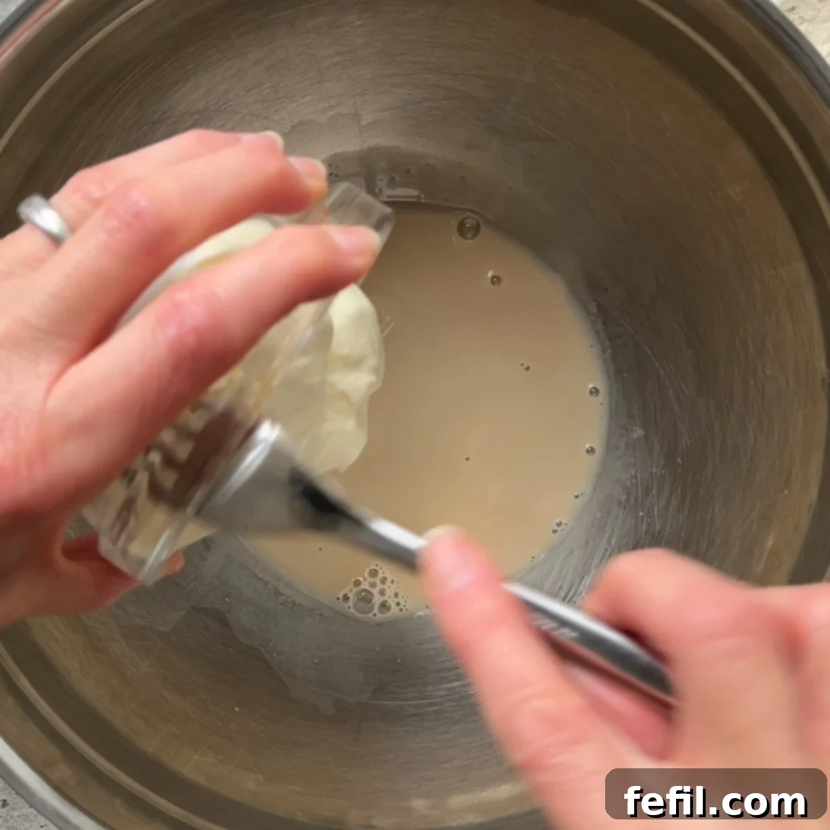 Hands adding a Greek yogurt from a small bowl into a metal mixing bowl containing water and yeast.