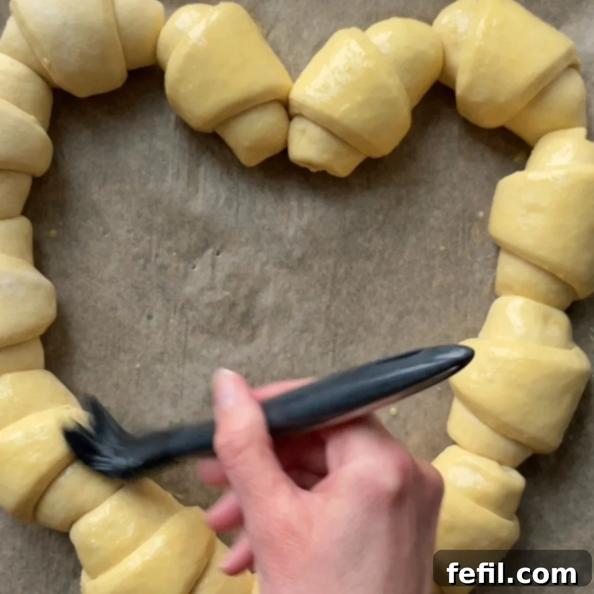 A hand uses a brush to apply egg wash to unbaked sausage rolls arranged in a heart shape on parchment paper.