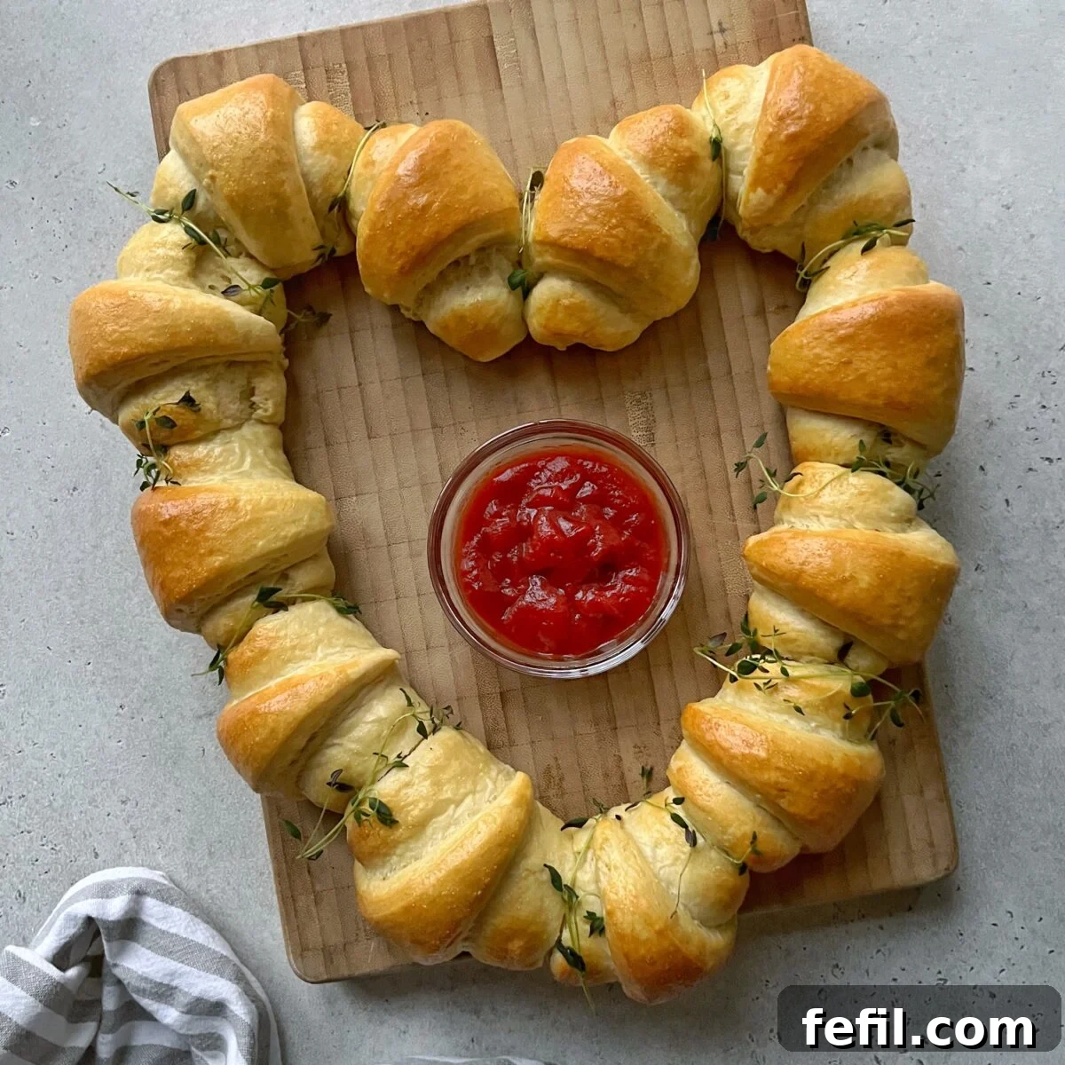 A heart-shaped ring of baked sausage rolls on a wooden board surrounds a bowl of ketchup.