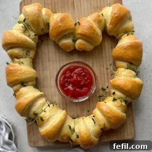 A heart shape of baked sausage rolls arranged around a glass bowl of ketchup on a wooden board, garnished with fresh herbs.