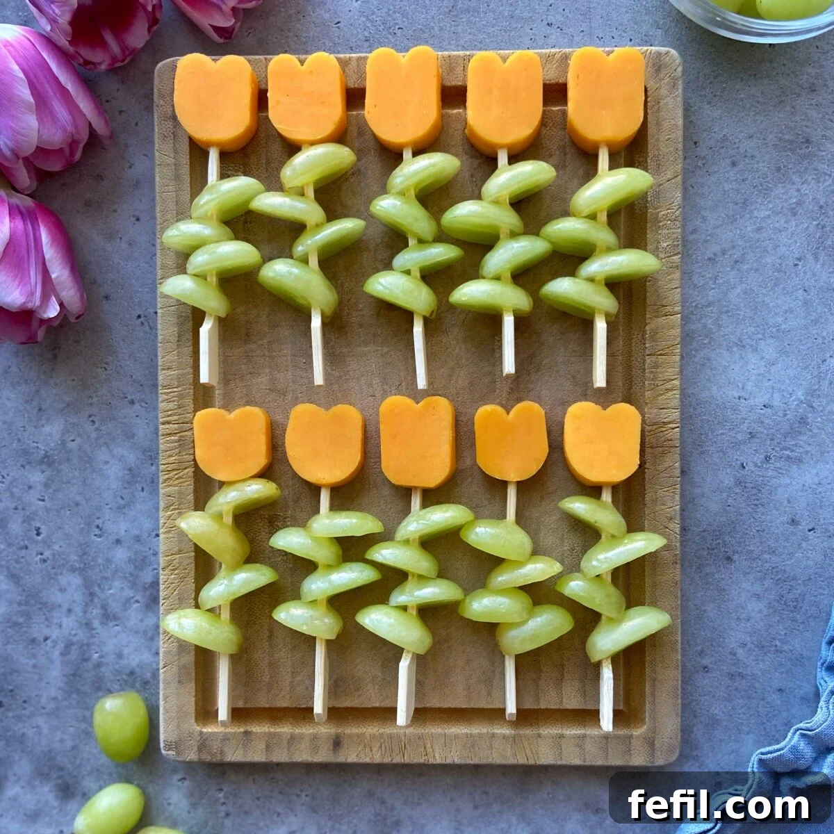 A rustic wooden board features beautifully arranged Cheese and Grape Skewers, with vibrant red cheddar cheese meticulously cut into tulip shapes, elegantly paired with fresh green grapes designed to resemble flower stems. The arrangement creates a visually appealing and fresh appetizer.