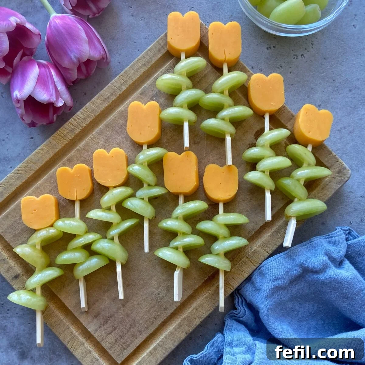 An inviting close-up of colorful Cheese and Grape Skewers artfully displayed on a wooden board. The skewers feature bright green grapes and charming tulip-shaped red cheddar cheese, complemented by soft pink tulips and a bowl of fresh grapes nearby, setting a lovely, fresh scene.