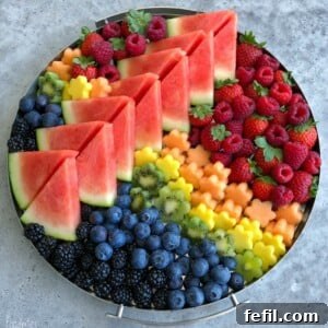 A round tray filled with a vibrant fruit medley—neatly arranged slices of watermelon, piles of raspberries, strawberries, blackberries, blueberries, kiwi, and flower-shaped pieces of cantaloupe and honeydew melon.