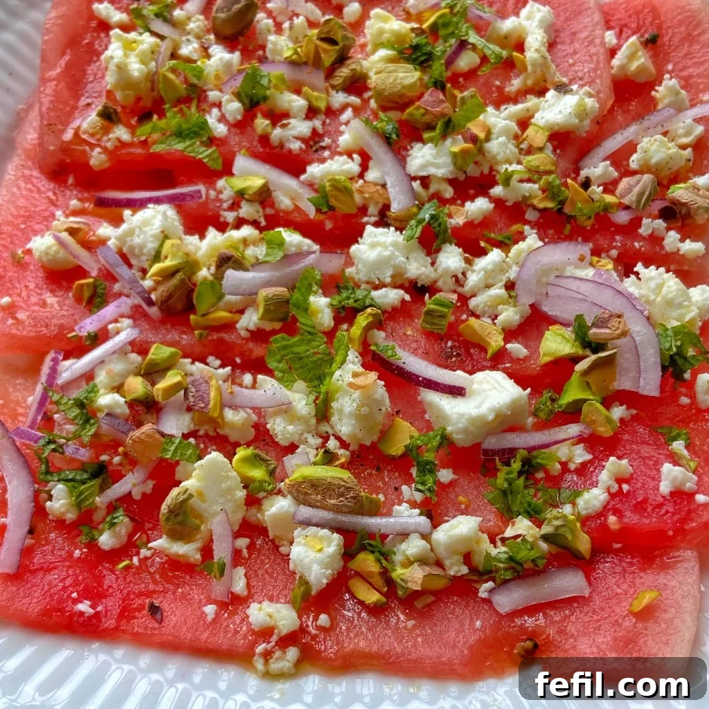 Thin slices of watermelon feta carpaccio are elegantly topped with crumbled feta cheese, finely diced red onion, toasted chopped pistachios, and fresh mint leaves, all artfully arranged on a pristine white plate, ready to be served.