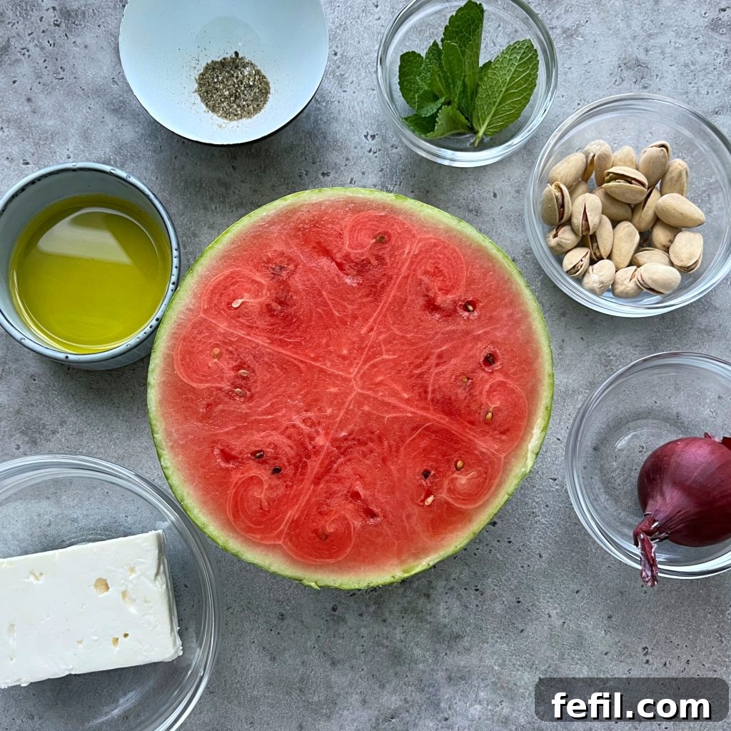 Close-up view of the ingredients for watermelon feta carpaccio arranged neatly on a wooden board, including a block of feta, a red onion, a pile of pistachios, fresh mint, and a bottle of olive oil.