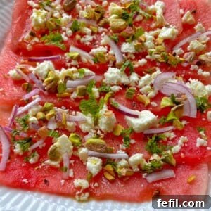 Thin slices of watermelon feta carpaccio are topped with crumbled feta, red onion, chopped pistachios, and fresh mint, all beautifully arranged on a white plate.