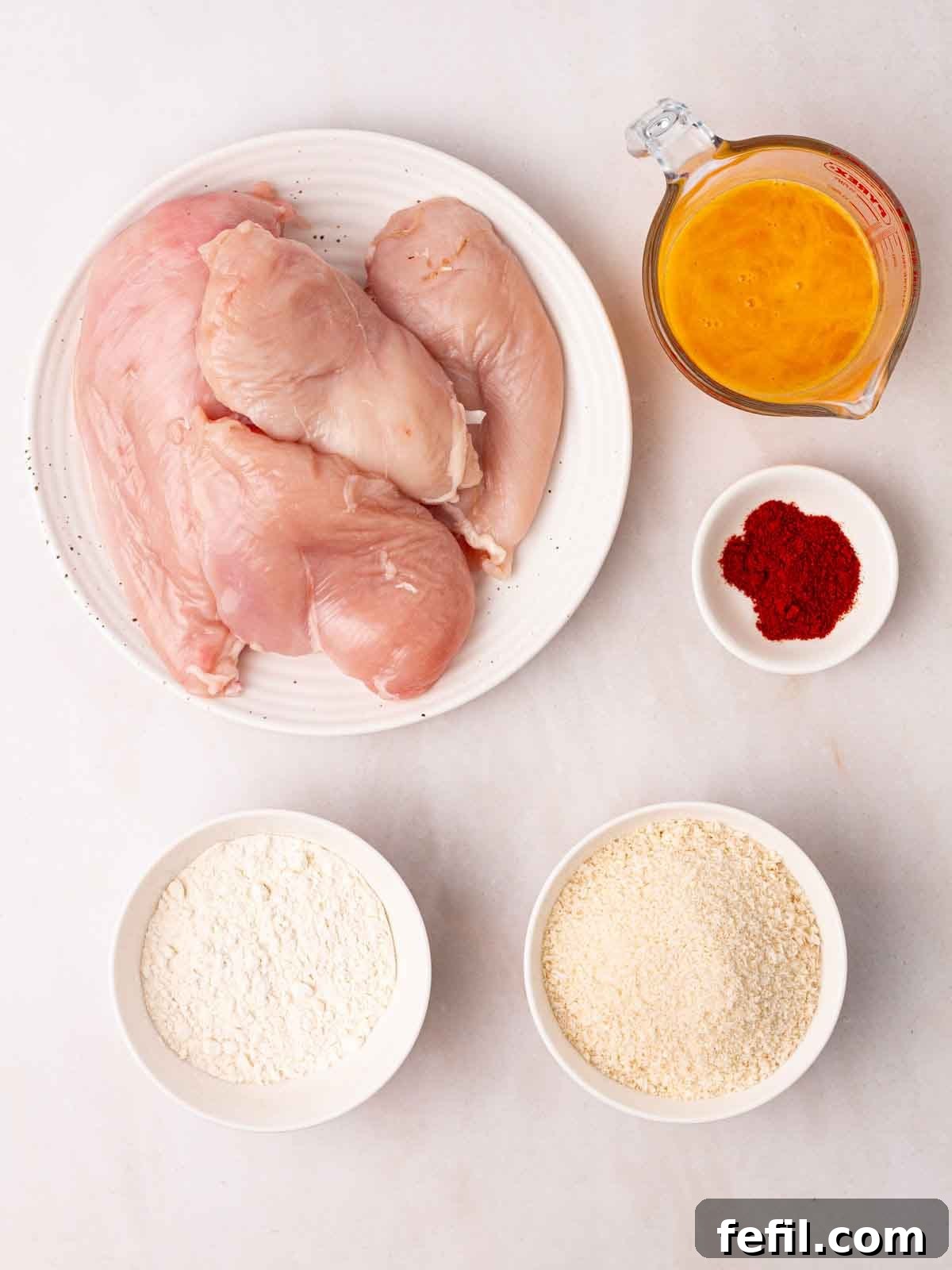 Raw ingredients for chicken goujons neatly laid out on a clean white countertop, including fresh chicken breast, bowls of flour, paprika, beaten eggs, and breadcrumbs, ready for preparation.