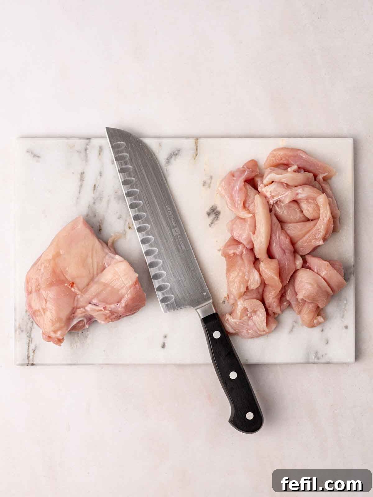 A marble chopping board with a sharp knife and raw chicken breast, clearly showing pieces already cut into uniform strips, demonstrating the initial prep stage.
