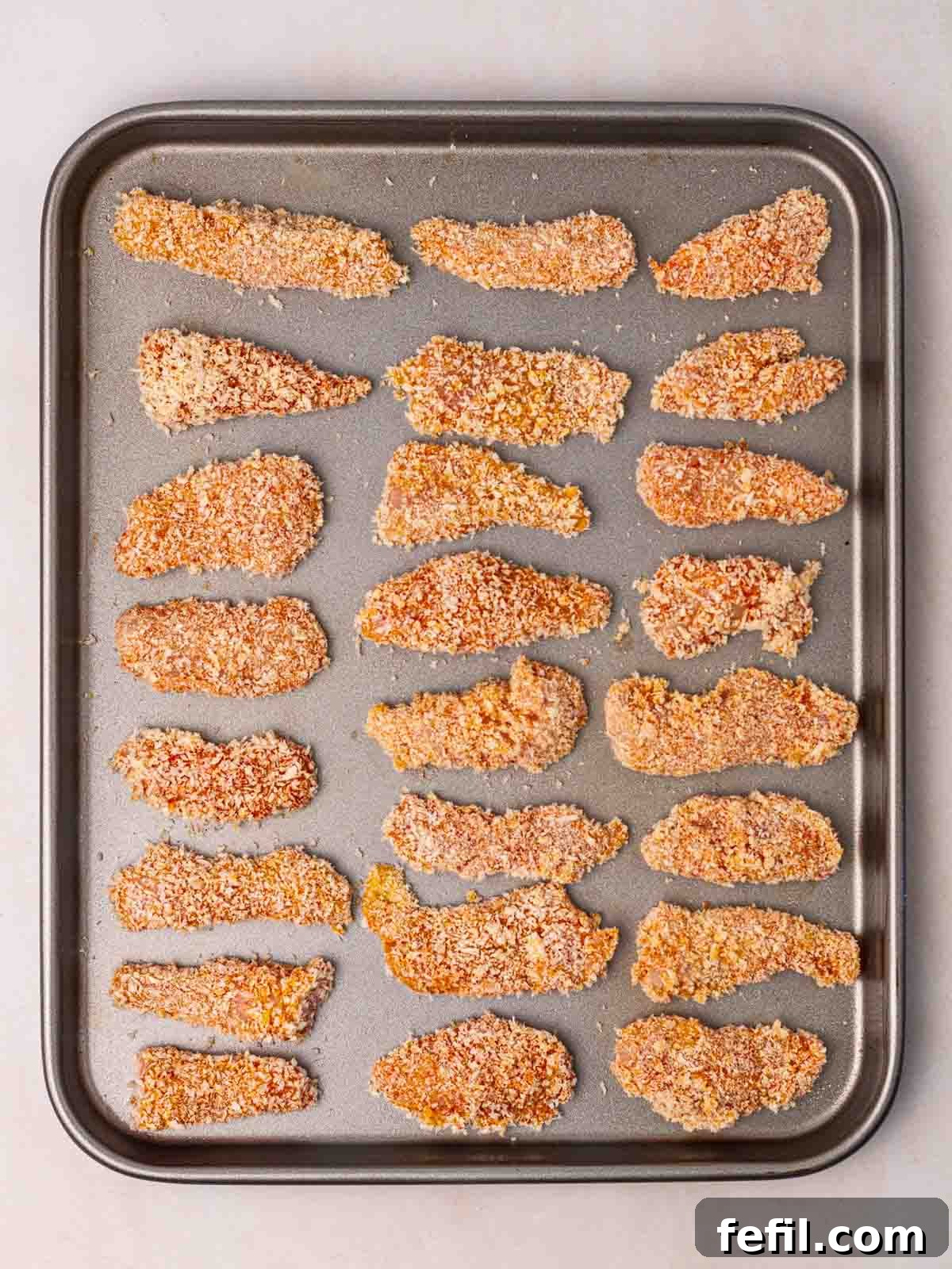 Raw, breaded chicken goujons neatly arranged on a greased baking tray, ready to be placed into the oven.