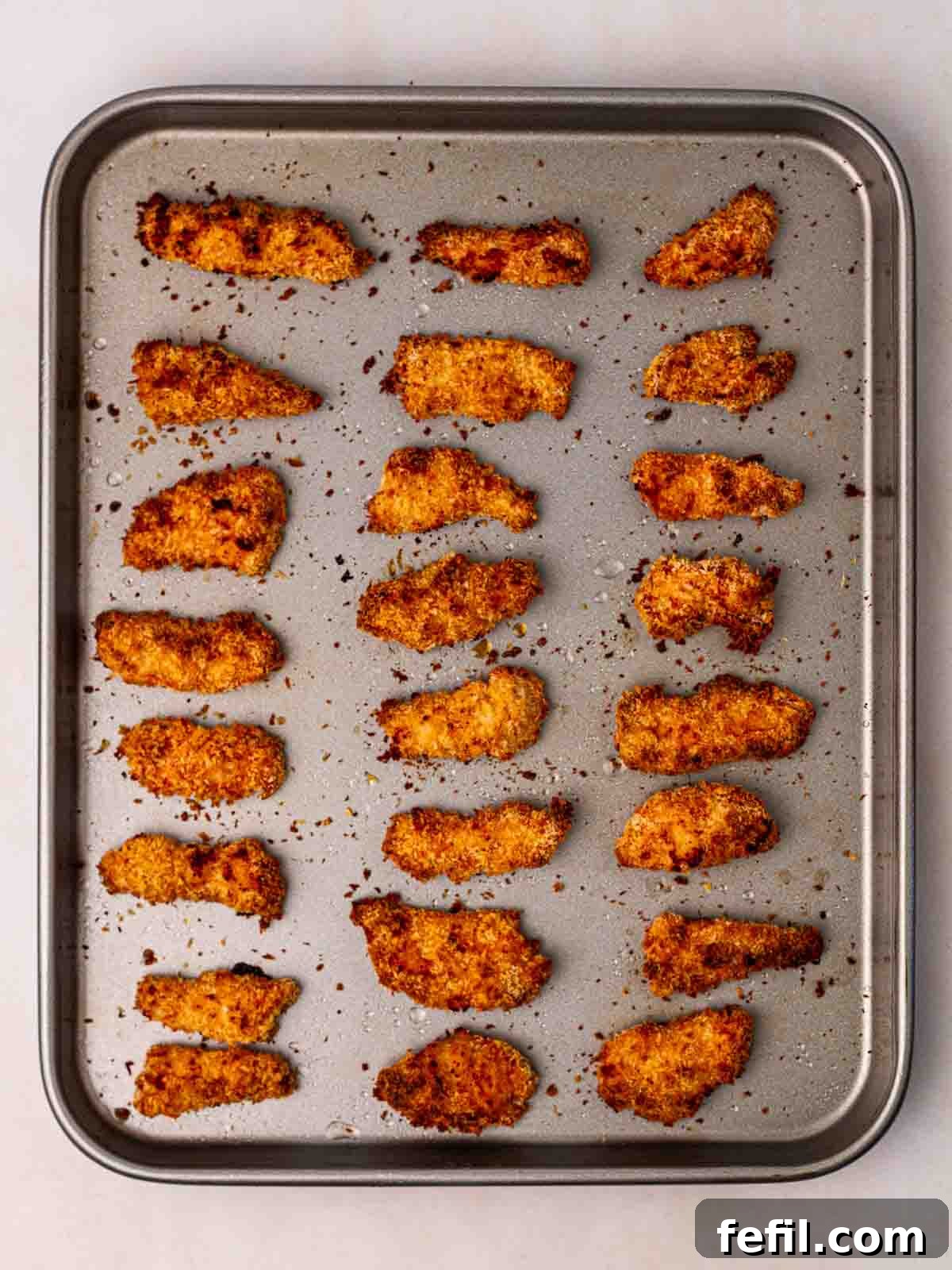 A baking tray filled with perfectly golden and crispy baked chicken goujons, just removed from the oven, emitting steam.