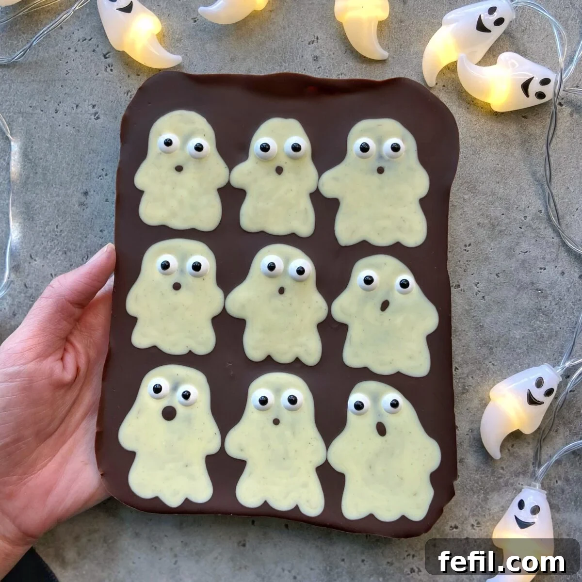 A hand holds a chocolate slab topped with nine white chocolate ghost bites, each with candy eyes, surrounded by ghost-shaped string lights on a gray surface.