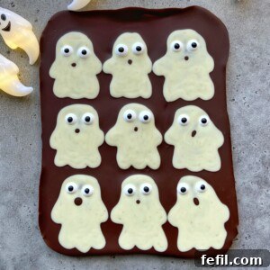 A slab of chocolate topped with nine white chocolate ghost shapes, each with candy eyes and different mouth expressions, arranged in a grid on a gray surface—perfect for fun food recipes or festive bites.