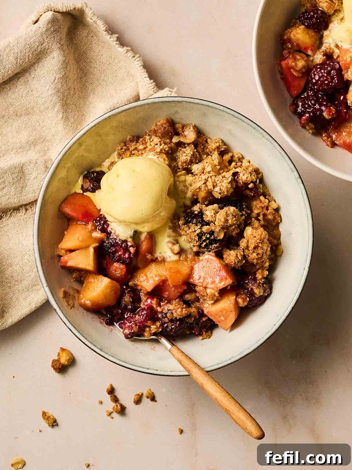 A delightful bowl of Apple and Blackberry Crumble, generously topped with a scoop of creamy vanilla ice cream, ready to be enjoyed.
