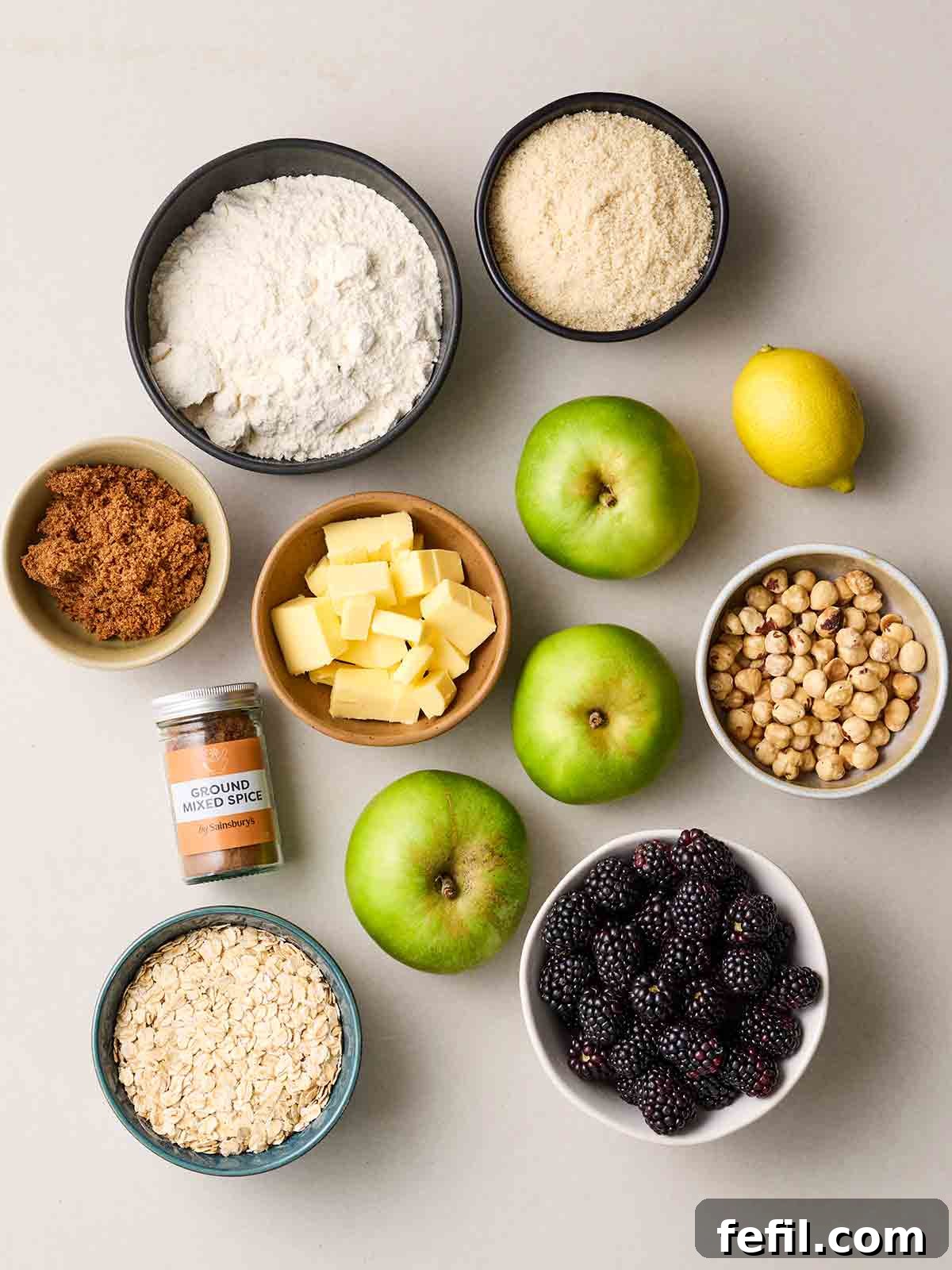 Fresh ingredients for Apple and Blackberry Crumble laid out on a clean white counter, including vibrant apples, dark blackberries, flour, lemon, oats, butter, ground mixed spice, brown sugar, and various nuts.