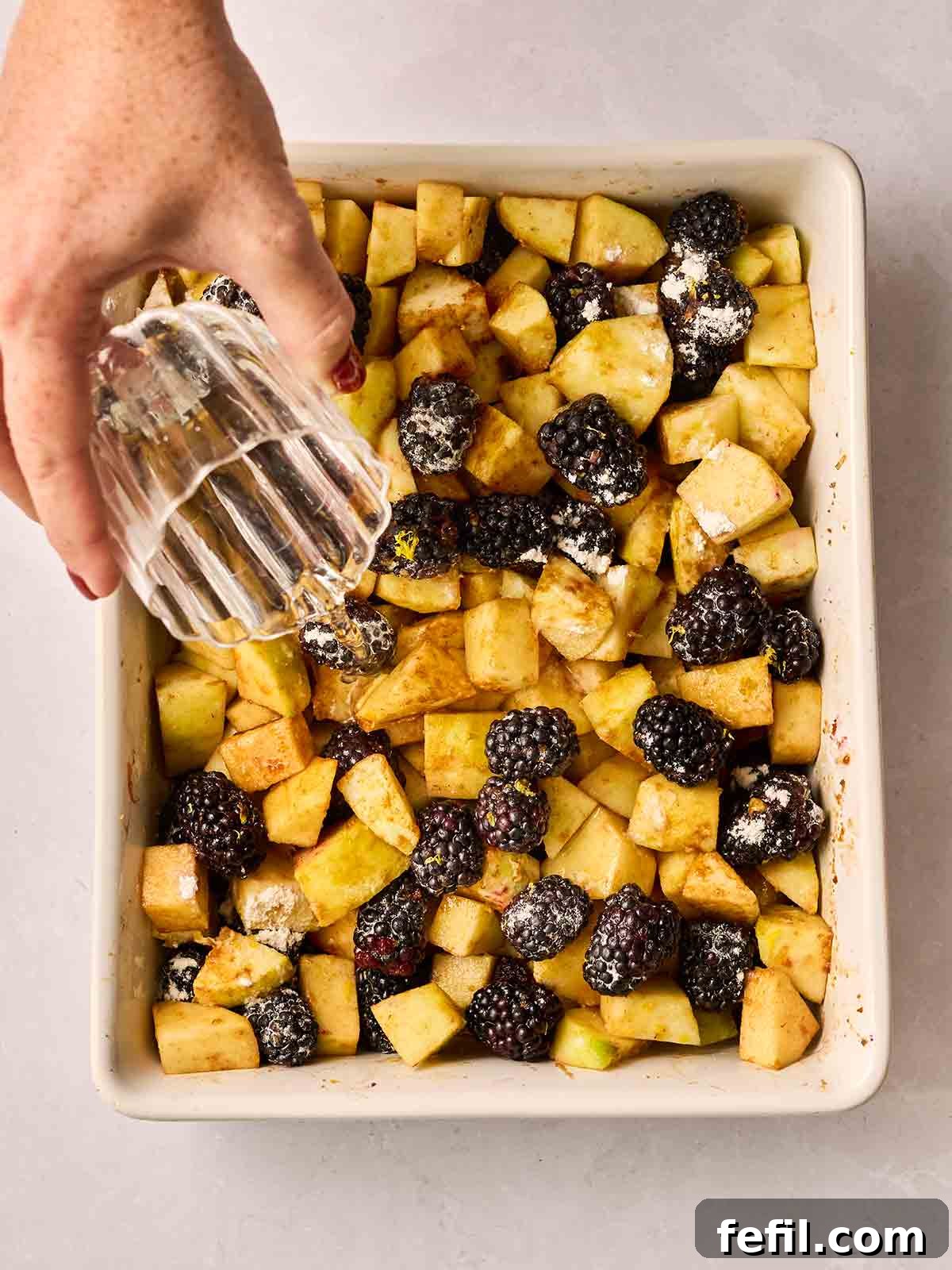 A hand is carefully pouring a stream of water from a small glass over the mixture of apples and blackberries in the ovenproof dish.