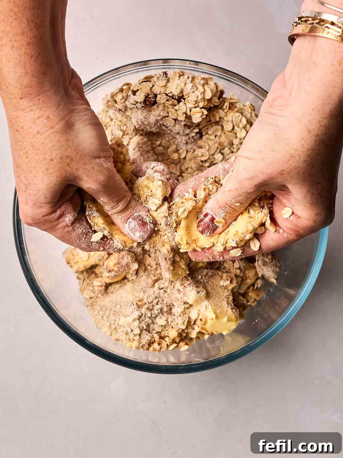 Two hands are actively rubbing together butter and dry ingredients in a large glass bowl, creating the crumbly topping for the dessert.