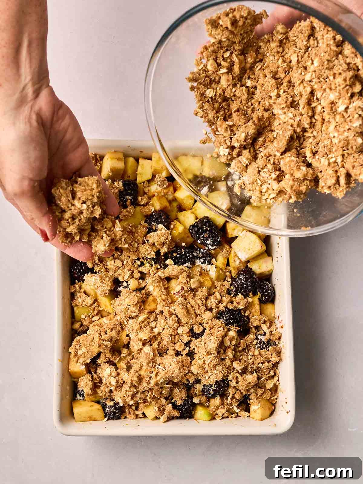 A generous layer of crumble topping is being scattered evenly over the apple and blackberry filling in a baking dish by hands from a glass bowl.